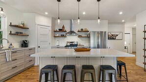 A modern kitchen with a large island, bar stools, pendant lights, and wooden shelves. - The Lake Hut - a perfect place to relax and play on shore of Lake Whatcom (Sedro Woolley)