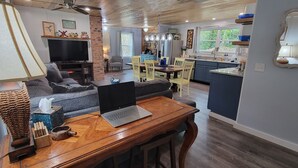 A modern living room with a sofa, a wooden table with a laptop, and a television. There is a kitchen area with a dining table and chairs, and a large window providing natural light.