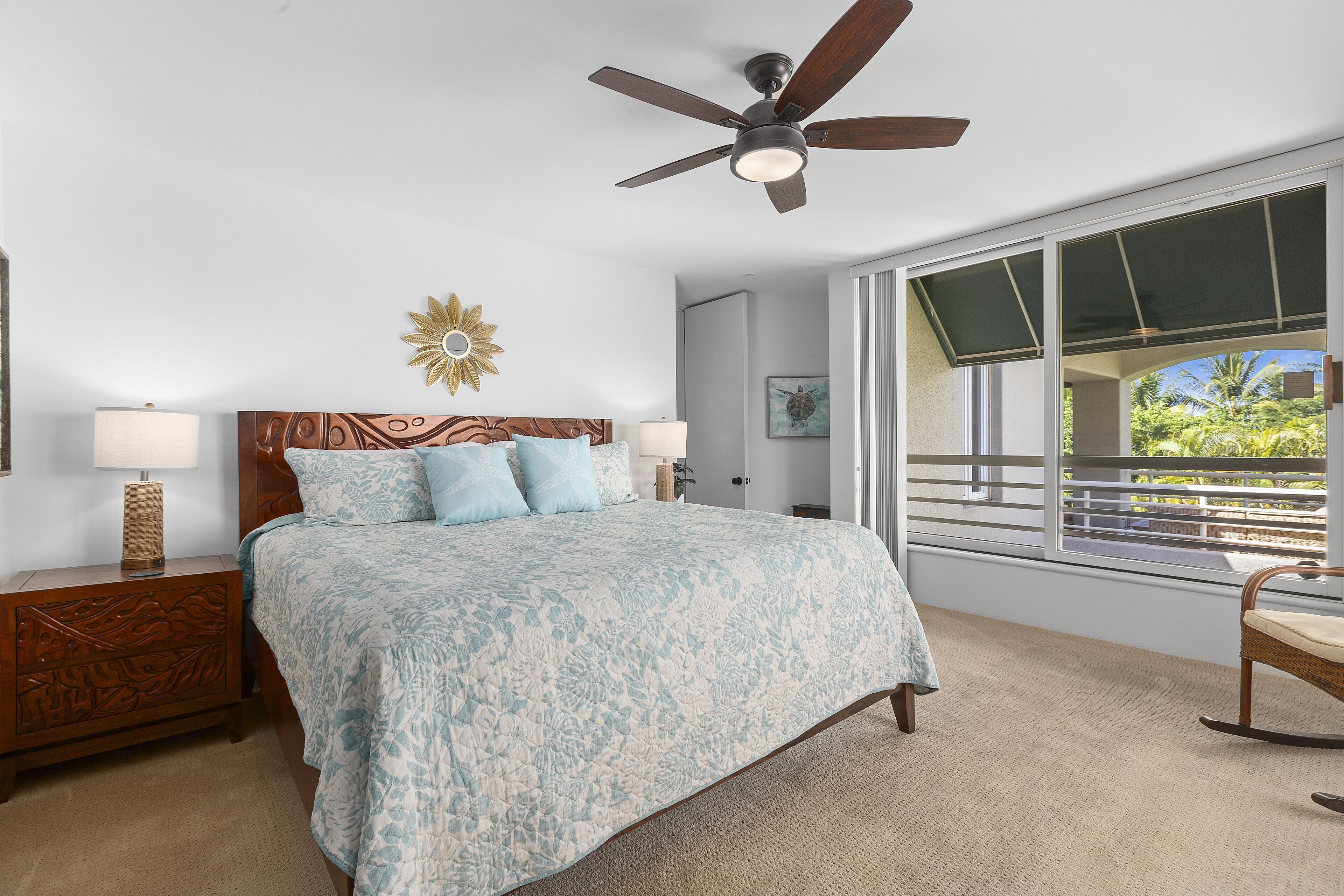 A bedroom with a bed, nightstands, a ceiling fan, a window with a view, and a decorative wall piece.