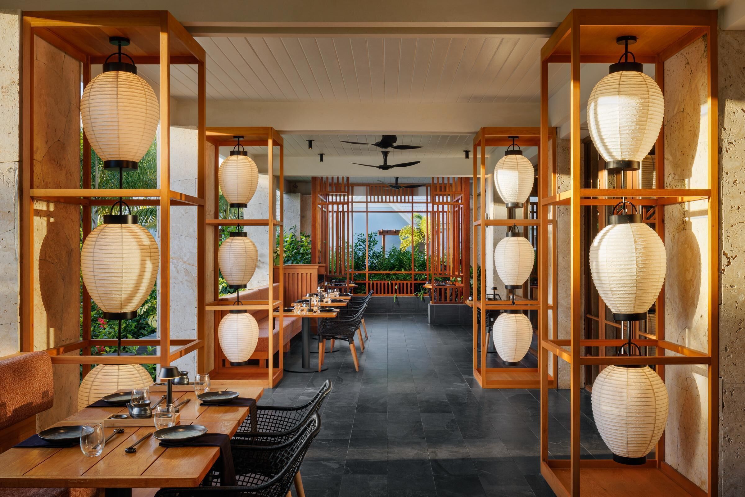 A restaurant with wooden tables and chairs, paper lanterns, and a view of greenery.