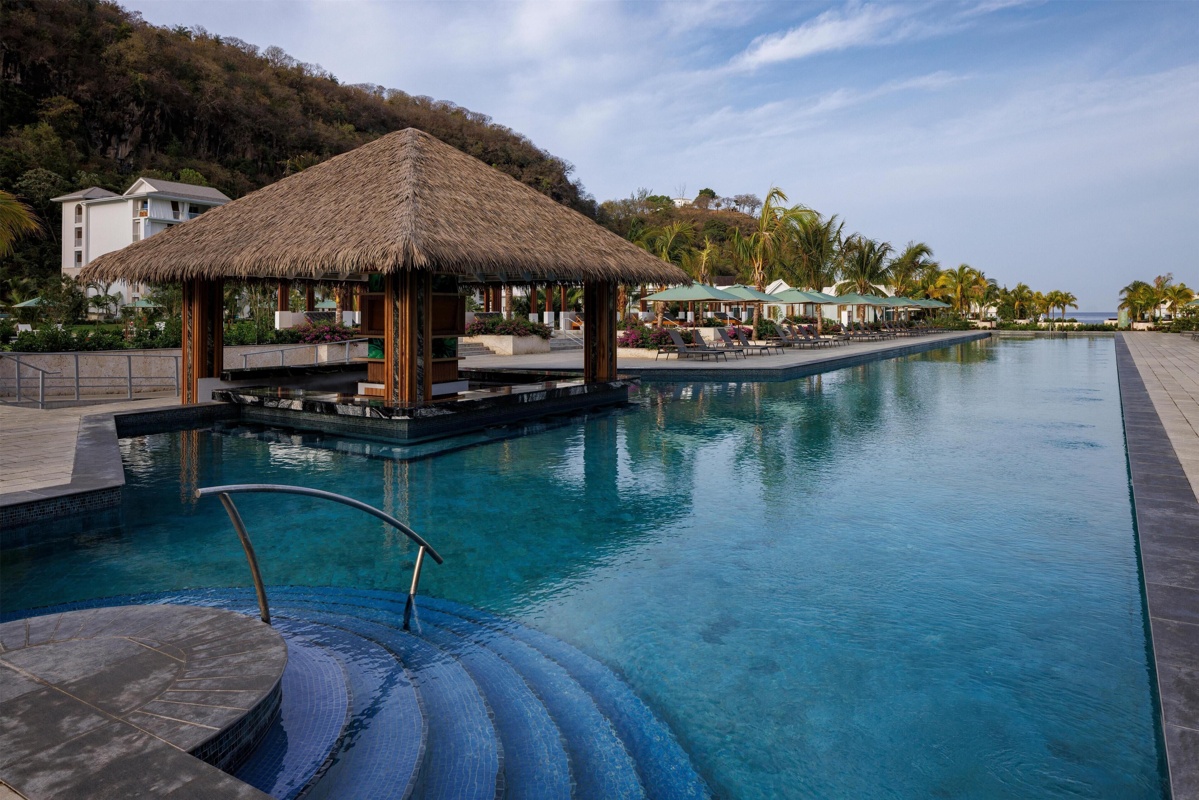 Une piscine d’hôtel avec un gazebo au toit de chaume, des chaises longues et une vue sur l’océan.