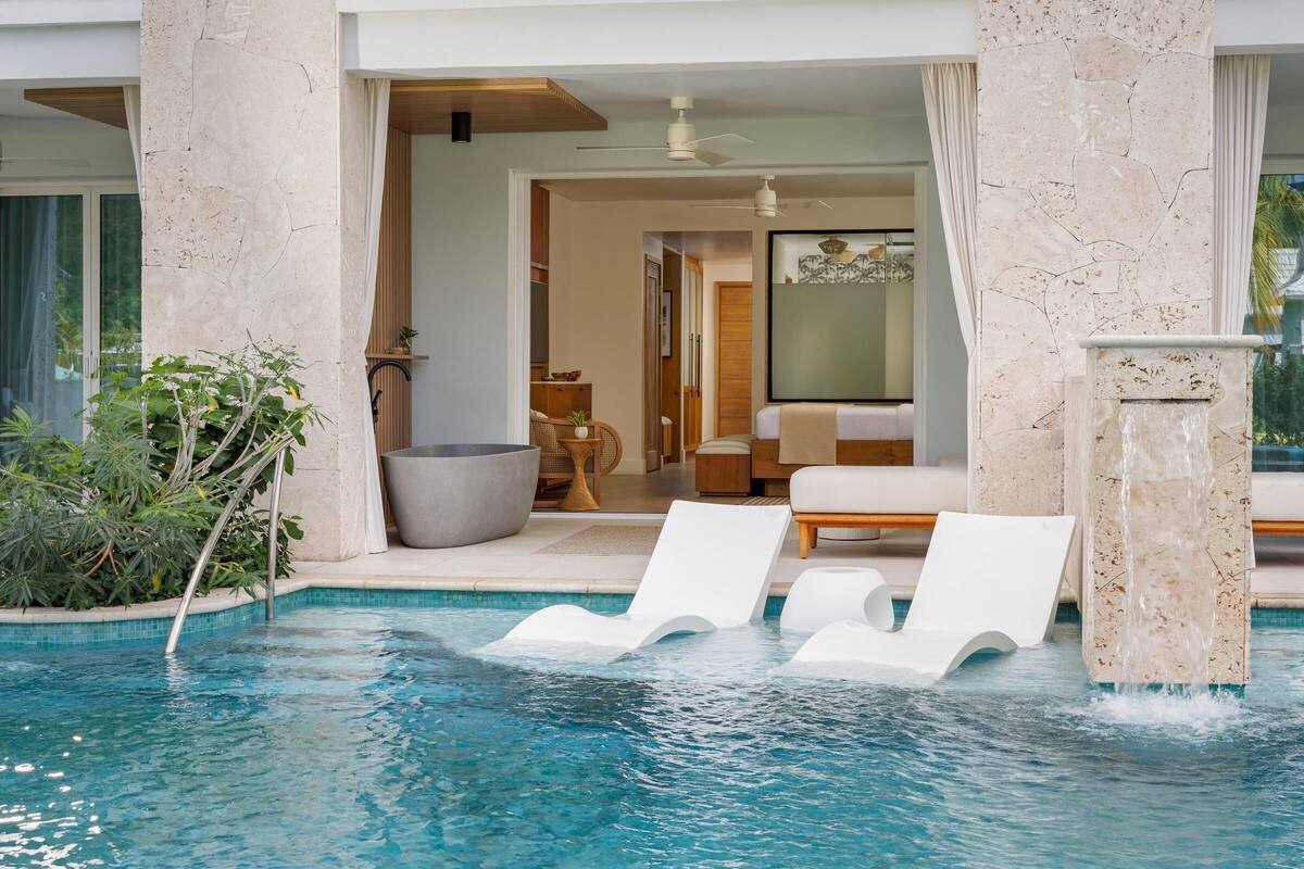 A pool area with white lounge chairs, a stone pillar, and a view into a room with a sofa and a dining table.