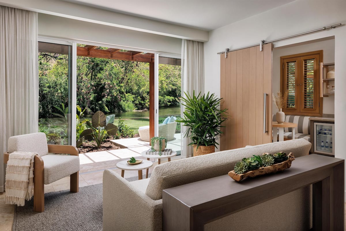 A modern living room with a sofa, armchair, and coffee table, a wooden cabinet, and a view of a garden through large sliding doors.