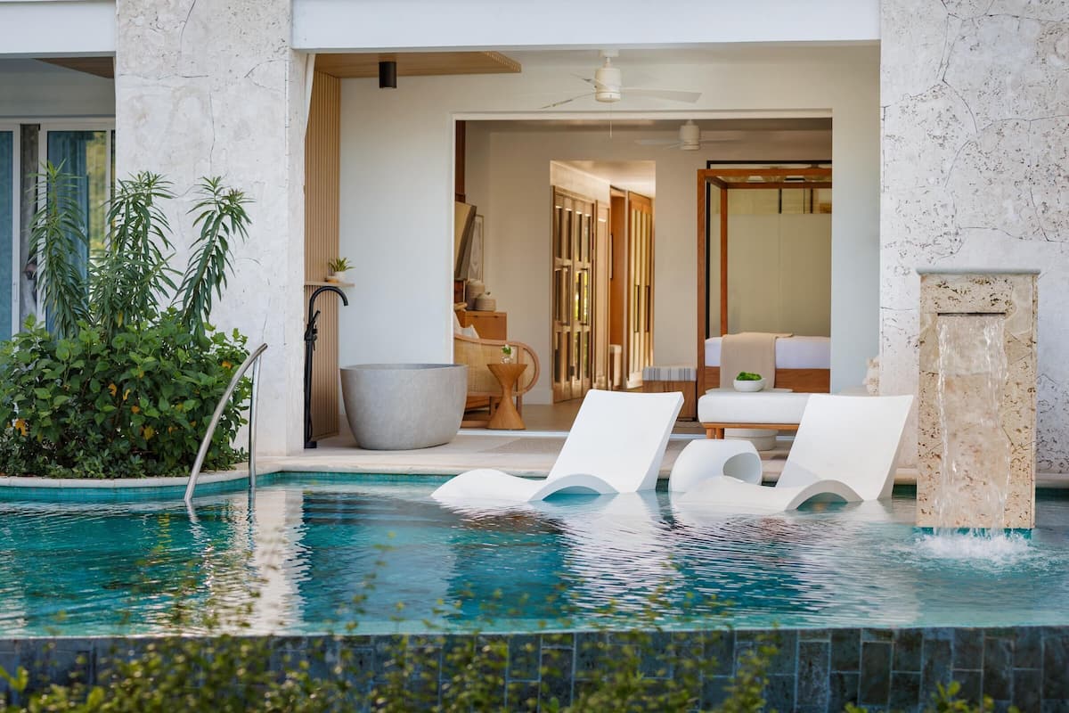 A modern pool area with two lounge chairs, a stone wall, and a view into a room with a bed and a ceiling fan.