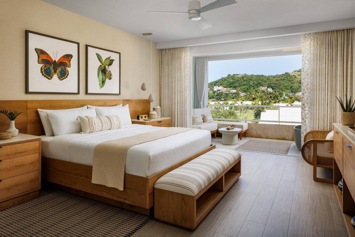 A spacious bedroom with a large bed, a wooden headboard, a bedside table, a bench, a nightstand, a window with a view of a green landscape, and a ceiling fan.