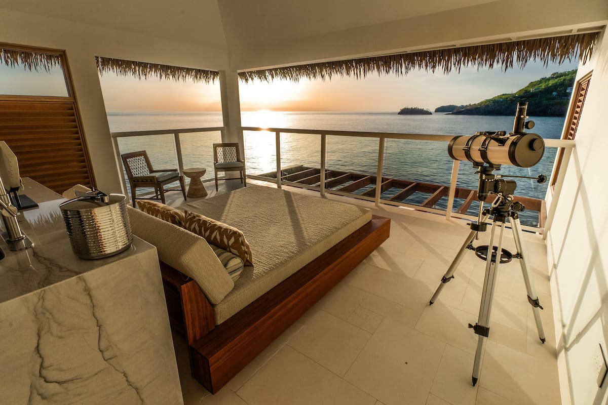 A bedroom with a large bed, a telescope, and a view of the sea at sunset.