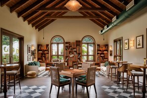 A spacious room with a high ceiling, wooden beams, and large windows. There are multiple seating areas with tables and chairs, and a large bookshelf filled with books and decorative items.