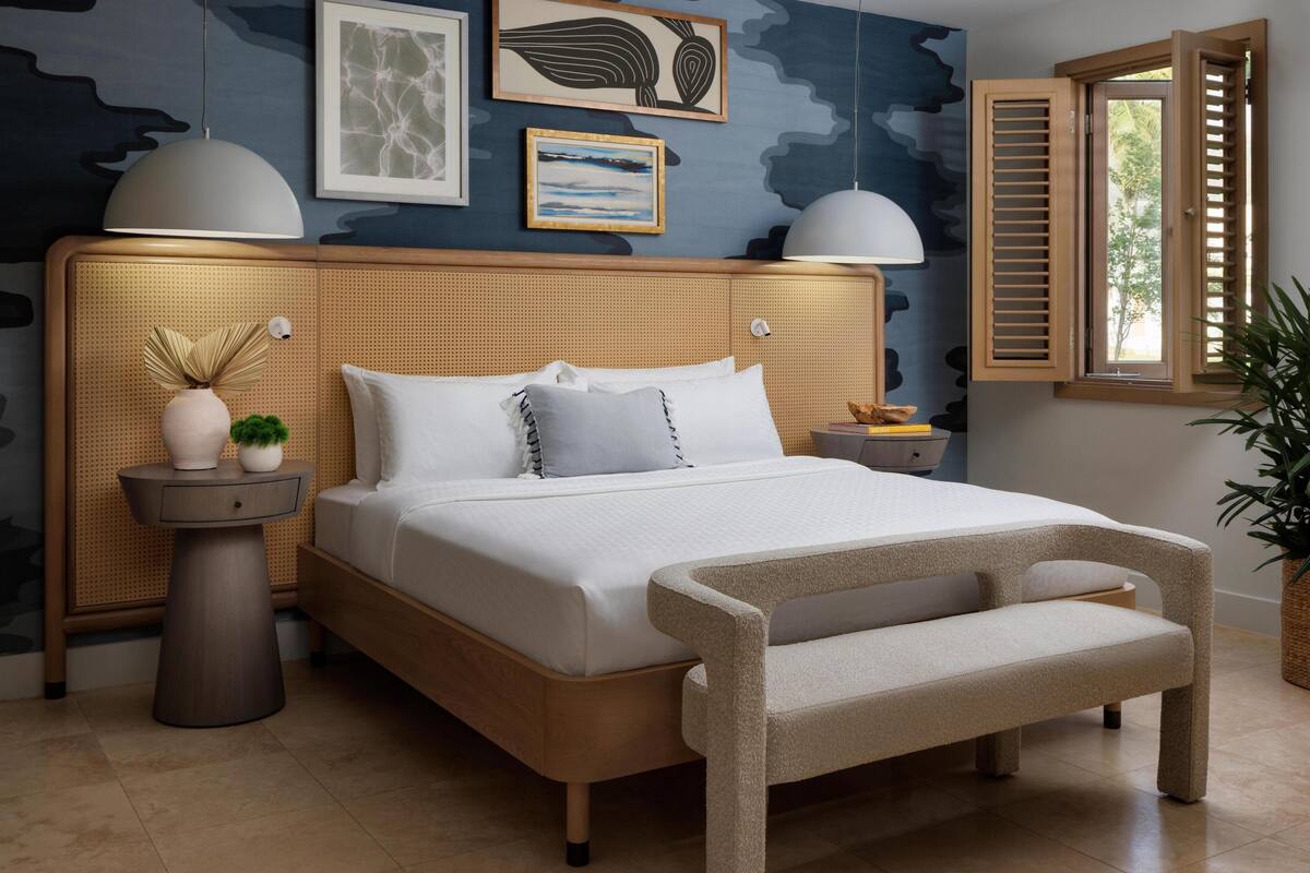 A modern bedroom with a rattan headboard, a large bed with white linens, two bedside lamps, and a window with wooden shutters.