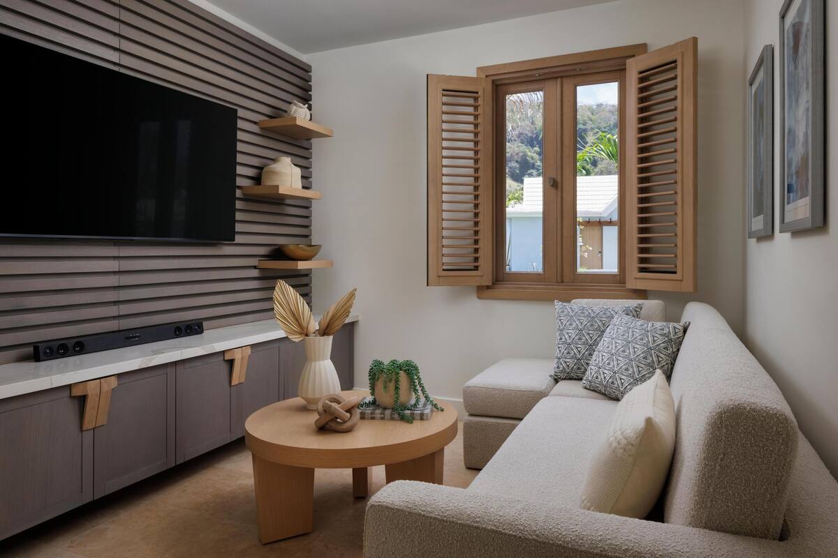 A modern living room with a flat-screen TV, wooden shelves, a round coffee table, a beige sofa, and a window with wooden shutters.