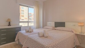 A hotel room with two beds, a window with a view of buildings, a desk with a lamp, and a dresser. - 080 - Gaviotas 004 - comfortHOLIDAYS (Santa Pola)
