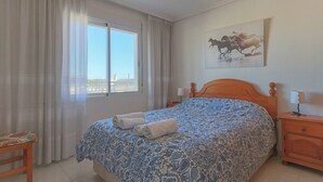 Een slaapkamer met een bed, gordijnen, een raam, een schilderij en een nachtkastje.