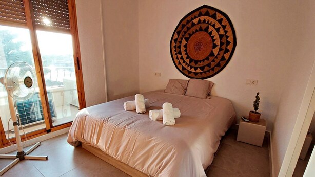 A bedroom with a large bed, a wall hanging, a standing fan, and a small table with a plant. - 082 - Gran Playa 005 - comfortHOLIDAYS (Santa Pola)