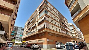 A multi-story residential building with balconies, a parking area in front, and a clear sky. - 084 - Isabel La Católica - comfortHOLIDAYS (Santa Pola)