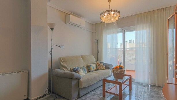 A living room with a sofa, a coffee table, a chandelier, and a view through a glass door. - 084 - Isabel La Católica - comfortHOLIDAYS (Santa Pola)