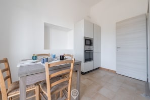 Een kleine keuken met een magnetron, oven en witte keukenkastjes. Er staat een eettafel met stoelen en een paar spullen op.