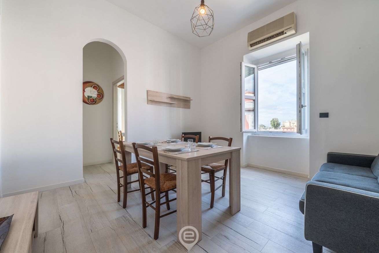 A dining area with a wooden table and chairs, a hanging light fixture, and a window with a view of buildings.