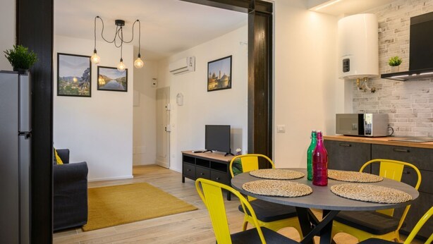 Una cocina moderna con un salpicadero de ladrillo, un comedor con sillas amarillas y un televisor sobre una mesa de madera.