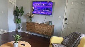 Ein Wohnzimmer mit einem gelben Sessel, einem Couchtisch aus Holz mit einer Pflanze, einem an der Wand montierten Fernseher und einem gemusterten Teppich.