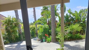 A view through a glass door of a room with a wicker chair, a potted plant, and a doormat. - Iconic Wainui Beach a 100m stroll. Relax in Tropical type surroundings. (Gisborne)