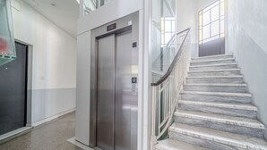 Un interior con un ascensor fijo, una escalera de mármol y una barandilla de cristal.