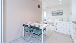 Un cuarto moderno y bien iluminado con una mesa de comedor dispuesta para cuatro personas, un sofá blanco y un cuadro de un paisaje urbano en la pared.