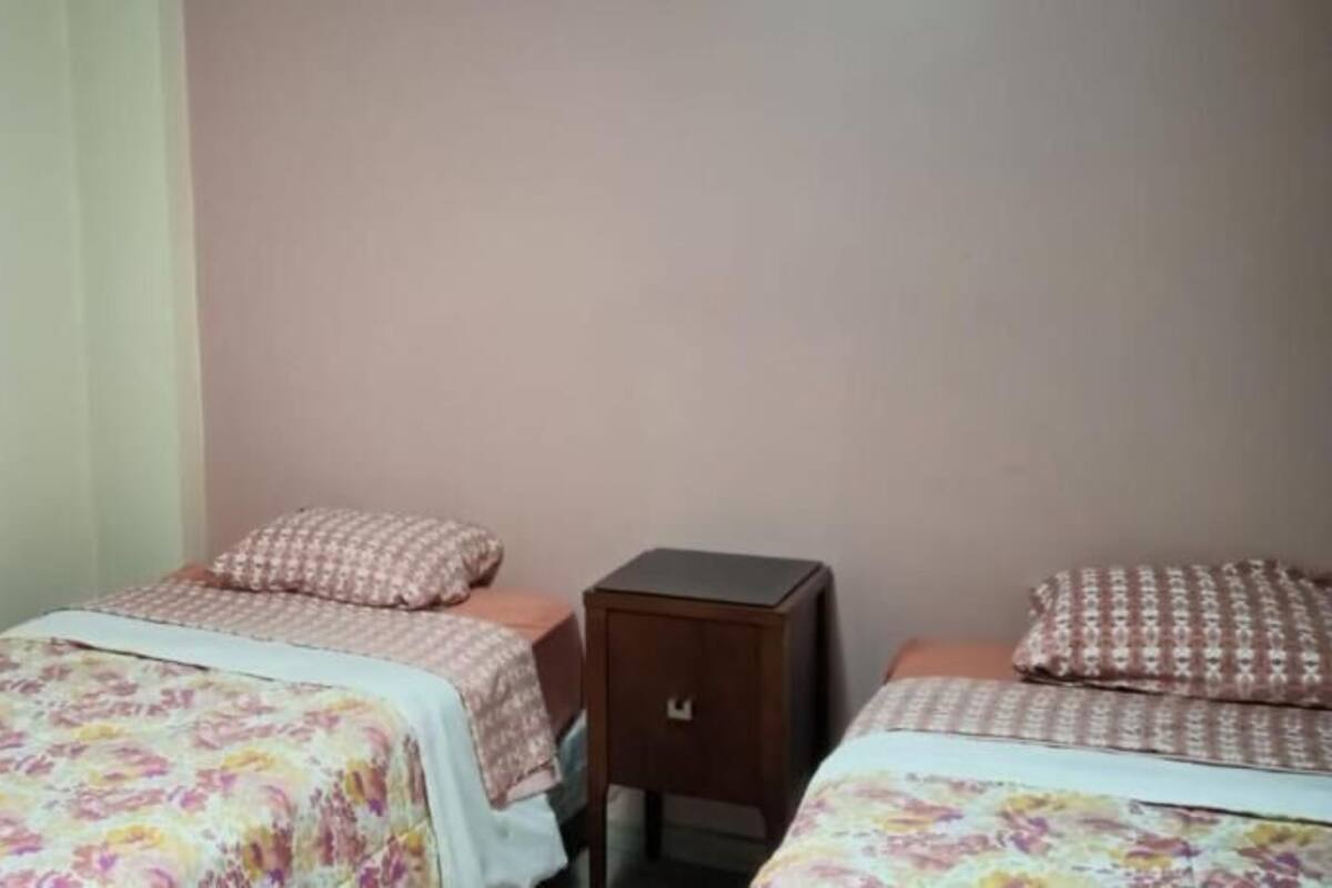 A room with two beds, each with a floral bedspread and patterned pillows. There is a small wooden nightstand between the beds.