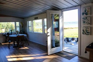 A room with a large sliding glass door leading to an outdoor patio, a dining area with a round table and chairs, and a view of a natural landscape. - Beautiful waterfront property (Chesterville)