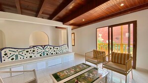 Una habitación con un sofá blanco, dos sillones de mimbre, una mesa de centro de vidrio con diseño floral y un amplio ventanal con vistas a un balcón.