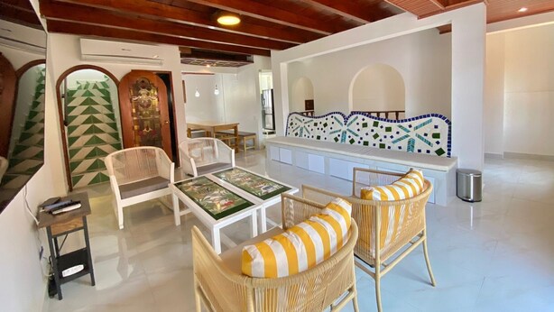 Amplia sala de estar con techo alto, paredes blancas y techo de madera. Hay sillas de mimbre con cojines a rayas amarillas y blancas, un sofá blanco y una pequeña mesa con camino de mesa verde.