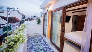 Un balcón con baldosas estampadas, una cama con cabecera de madera y vistas a edificios cercanos.