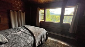 Un dormitorio de cabaña de madera con cama, cortinas y ventana con vistas a un bosque.