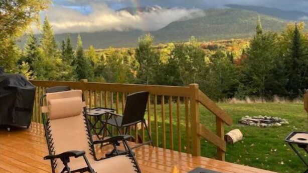 Una terraza con mobiliario exterior y un pozo de fuego, con vista a un paisaje montañoso y un arcoíris.