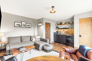 A modern living room with a grey sofa, a coffee table, and a leather couch. There's a kitchen area with wooden shelves and a black refrigerator.