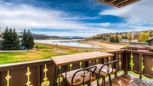 A wooden deck with a table and chairs overlooking a beach with boats, trees, and buildings in the background. - Lakeside One-Bedroom with Balcony/Views! Close to Keystone.  (Dillon)
