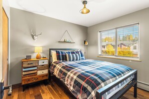 A bedroom with a bed, a dresser, a wall-mounted lamp, a window with blinds, and a ceiling light fixture.