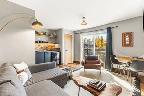 A modern living room with a grey sofa, a coffee table, and a dining area with a wooden table and chairs. - Lakeside One-Bedroom with Balcony/Views! Close to Keystone.  (Dillon)