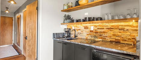 A modern kitchen with a stone backsplash, wooden shelves, and a black dishwasher.
