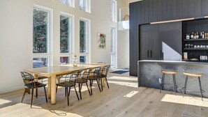 Moderni keittiö, jossa on baaritiski, suuret ikkunat ja ruokapöytä tuoleineen.