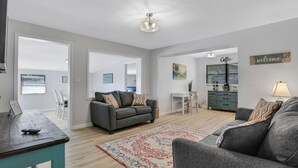 Een ruime woonkamer met een grijze bank, een salontafel en een tapijt met een patroon. Er is een eethoek met stoelen en een tafel, plus een kitchenette met een koffieapparaat.