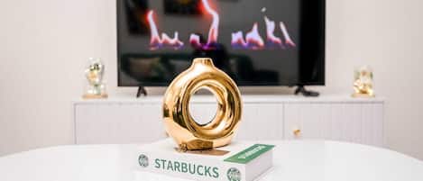 Una mesa blanca con una caja de Starbucks y un objeto dorado, un televisor al fondo y un armario blanco con adornos.