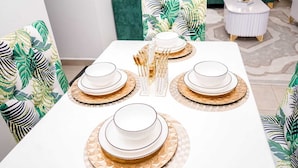 Una mesa de comedor con platos blancos, cuencos con detalles dorados y cubiertos con ribete dorado, rodeada de sillas con tapicería de estampado de hojas tropicales.