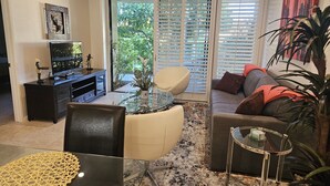 Un salon avec une table basse en verre, un canapé, un téléviseur et une porte-fenêtre coulissante donnant sur un jardin.