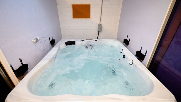 A hot tub with a rectangular shape and a white interior. There are four black jets visible on the side. The hot tub is placed against a light-colored brick wall with a wooden panel above it. - Sweet Home V - Family Cozy House, 20 minutes to downtown, 20 minutes to Airport (Houston)