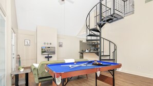 A living room with a pool table, a green sofa, a small table, and a spiral staircase.