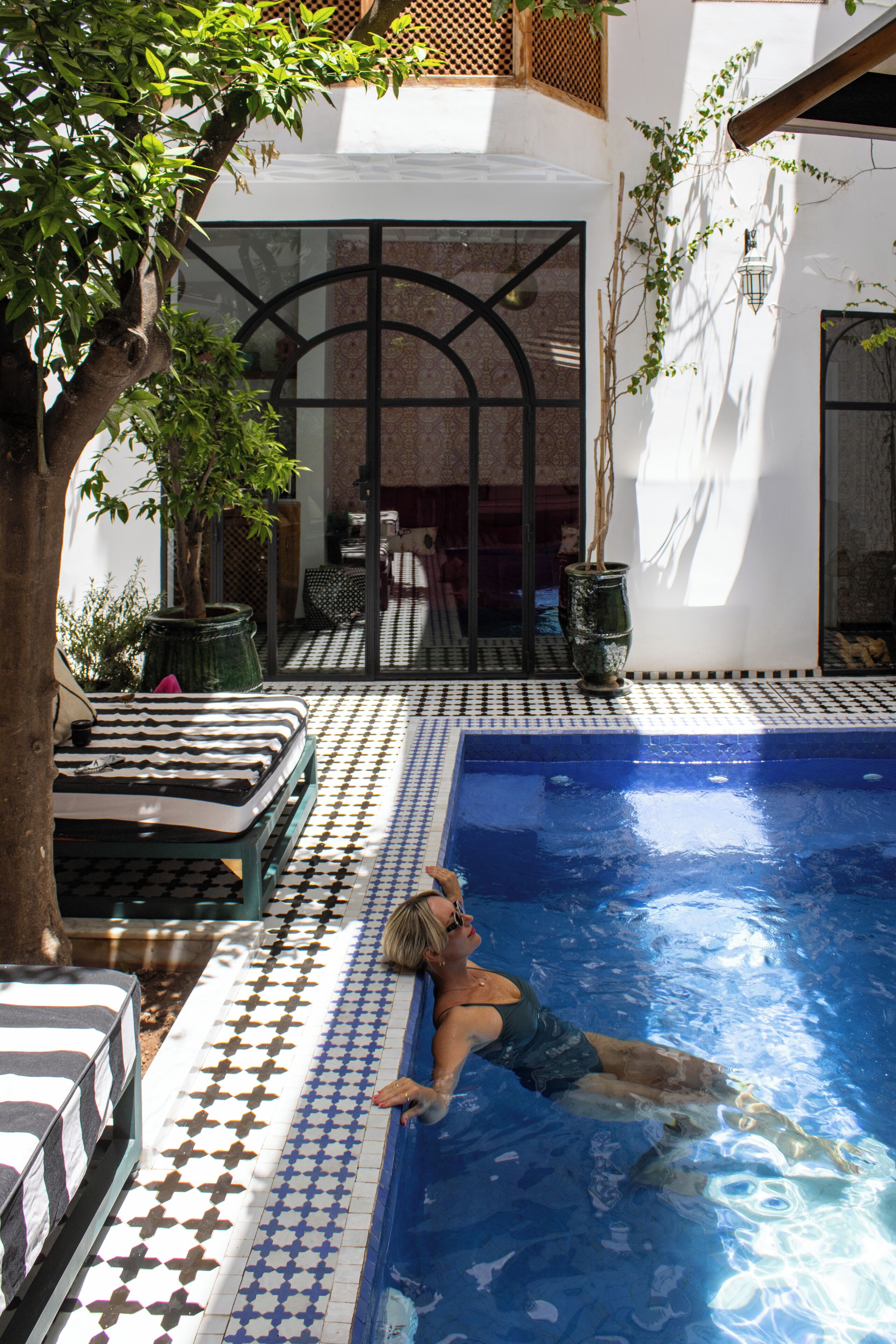 A swimming pool with a tiled floor, a lounge area with chairs and a table, and a tree providing shade.