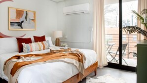 Une chambre moderne avec un grand lit, une table de chevet avec une lampe, un climatiseur fixé au mur et une vue sur le balcon extérieur.