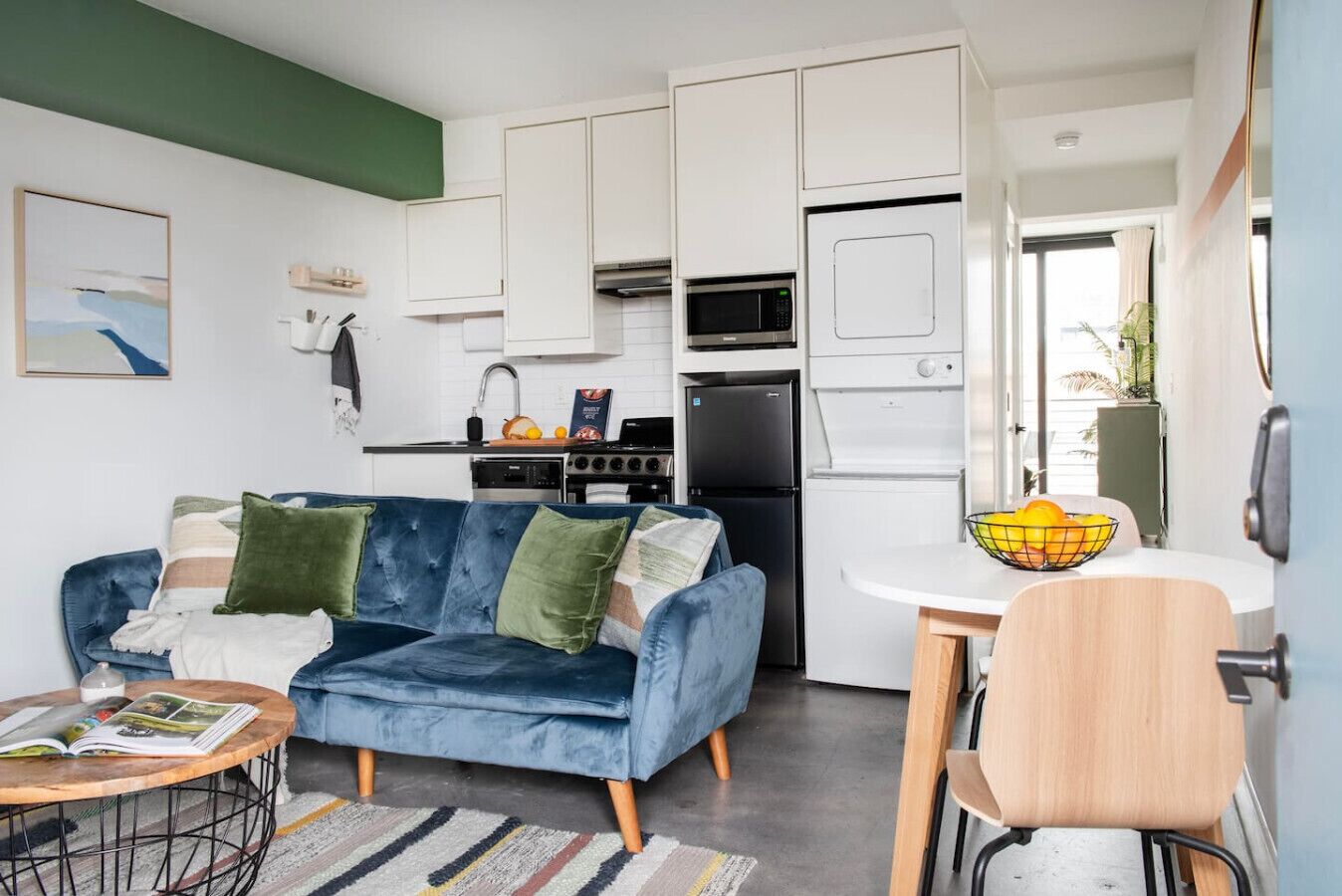 Un espacio habitable compacto con un sofá azul, una mesa de comedor con un cuenco de fruta y una kitchenette con microondas, estufa y refrigerador.