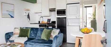 Un espacio habitable compacto con un sofá azul, una mesa de comedor con un cuenco de fruta y una kitchenette con microondas, estufa y refrigerador.