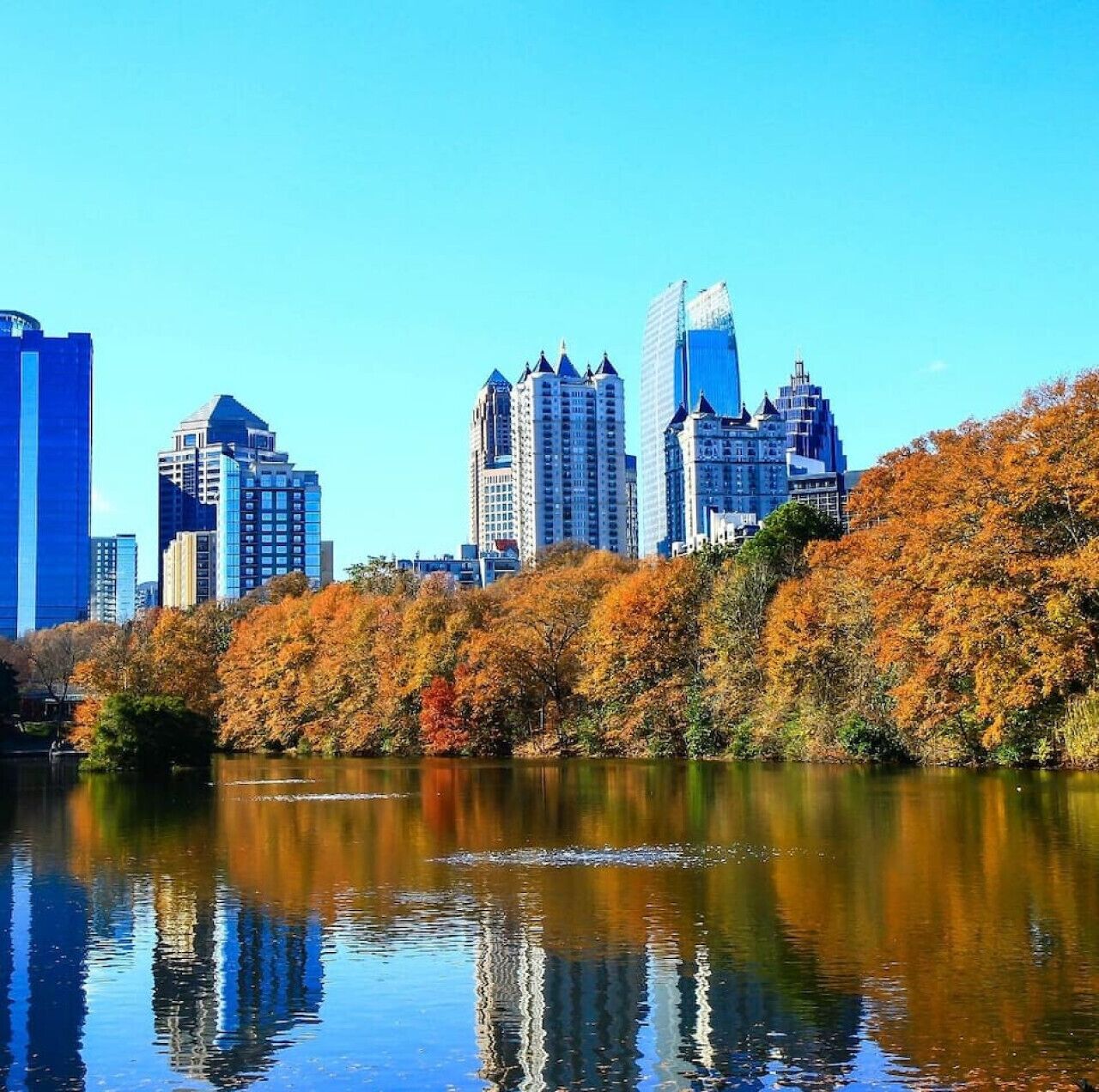 A city skyline with modern high-rise buildings and a calm lake reflecting the autumn foliage.