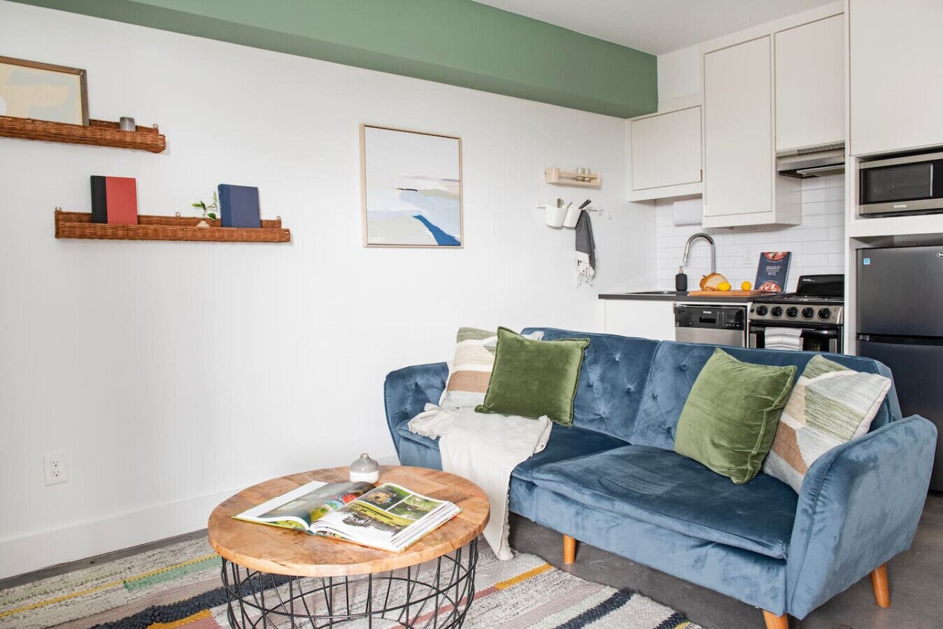 A living room with a blue velvet sofa, a round coffee table with magazines, a kitchen area with white cabinets and appliances, and a green accent wall.